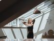 A woman standing in a sunlit attic, arms behind her head, exuding tranquility.