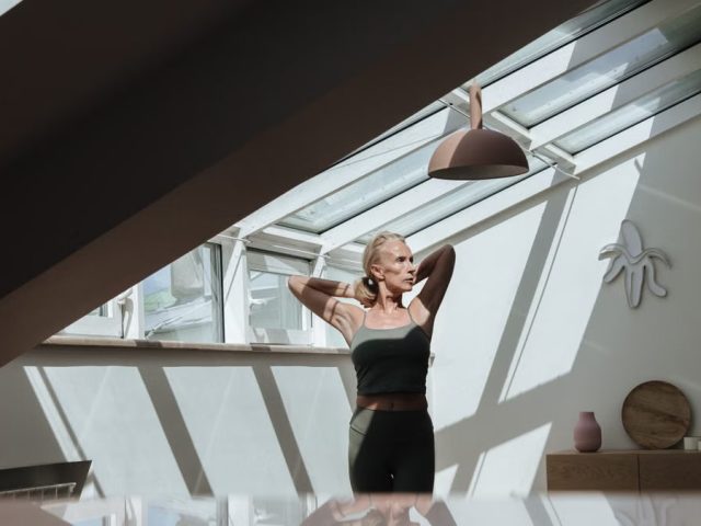 A woman standing in a sunlit attic, arms behind her head, exuding tranquility.